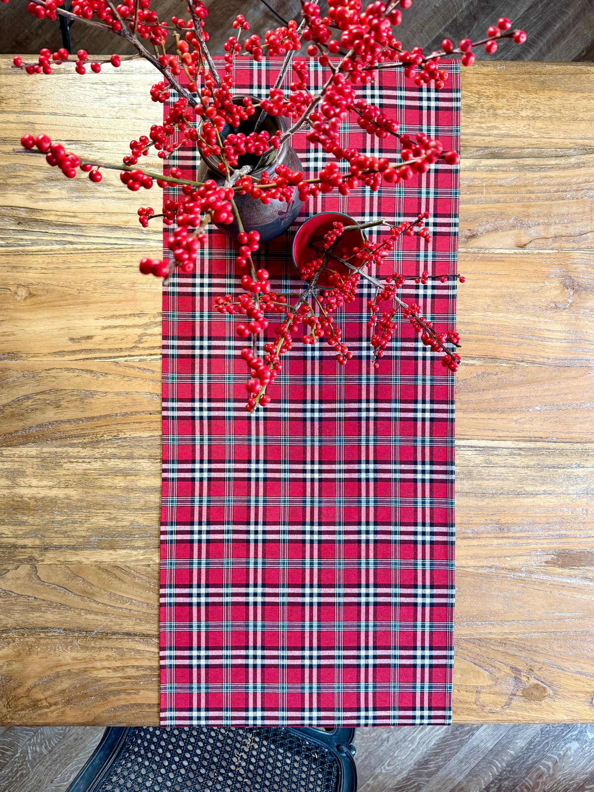Red tartan plaid Christmas table runner with navy blue and white checks, styled on a rustic wooden dining table with festive red berry decorations.